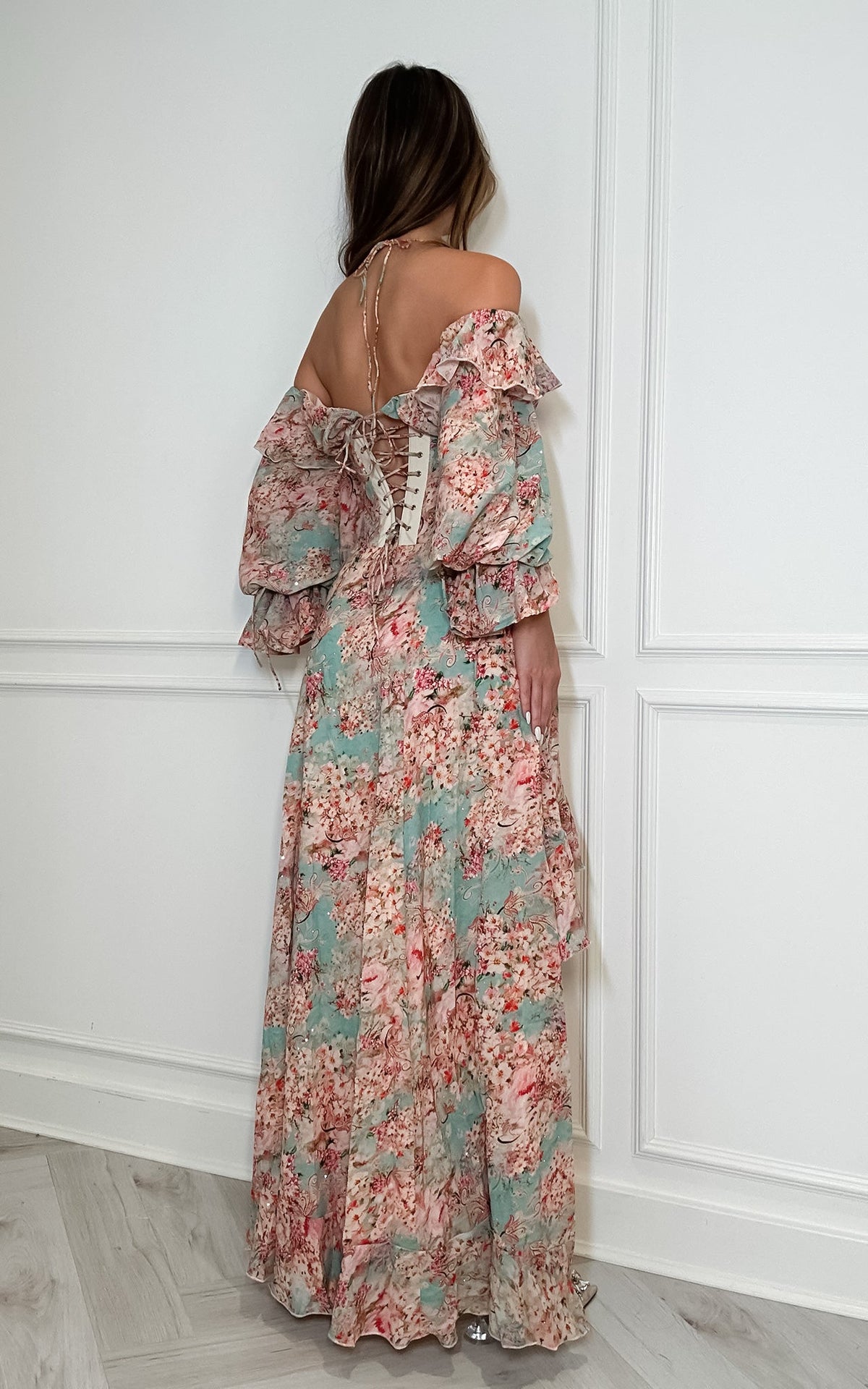 A woman viewed from behind wearing a long off-the-shoulder maxi dress with a ruffled lace-up back and a floral print in muted pink, sage green, and beige tones, standing against a white paneled wall.